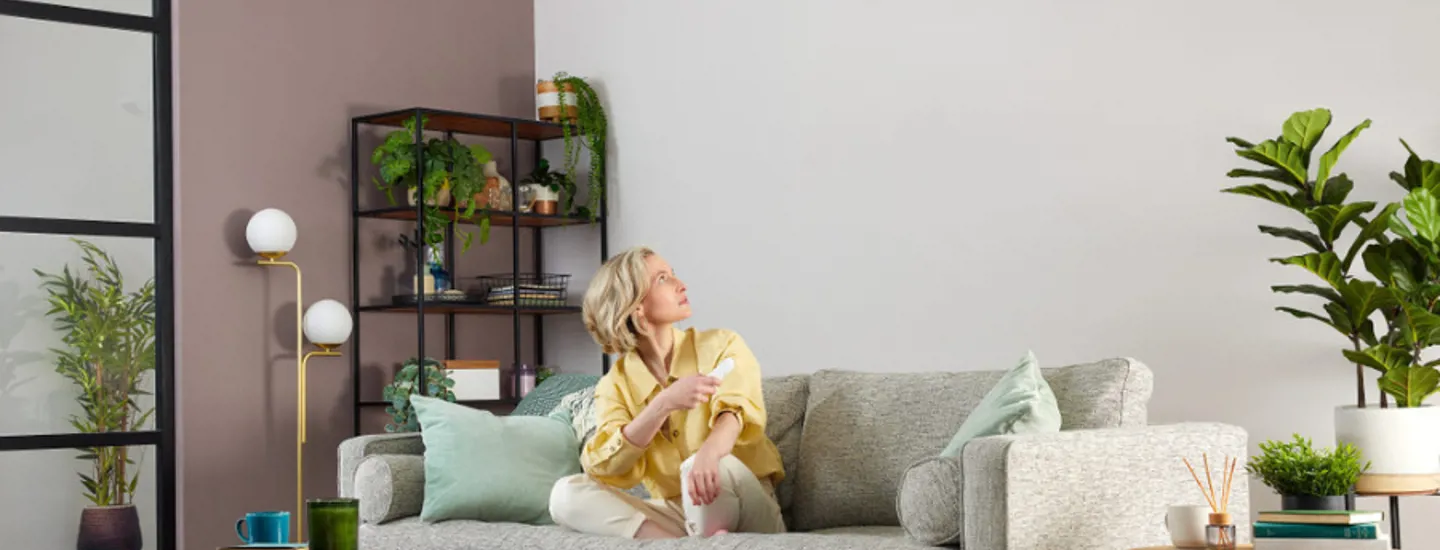 Kvinna sitter i soffa i vardagsrum och tittar upp mot en luftvärmepump på väggen